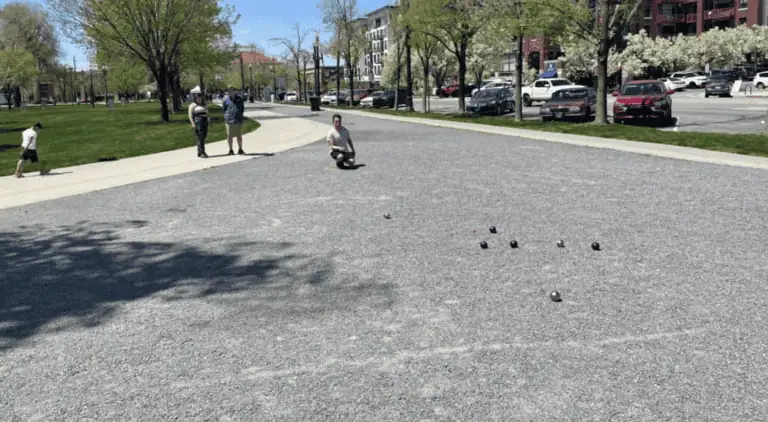People playing petanque on a gravel court in a city park.